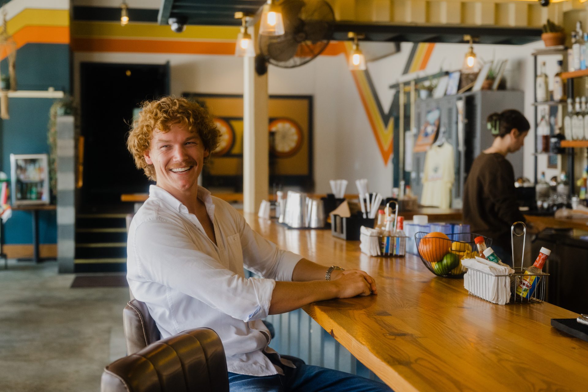 Young man with curly hair smiling confidently while leaning on a wooden desk in a creative office or co-working space for a lifestyle brand photography session