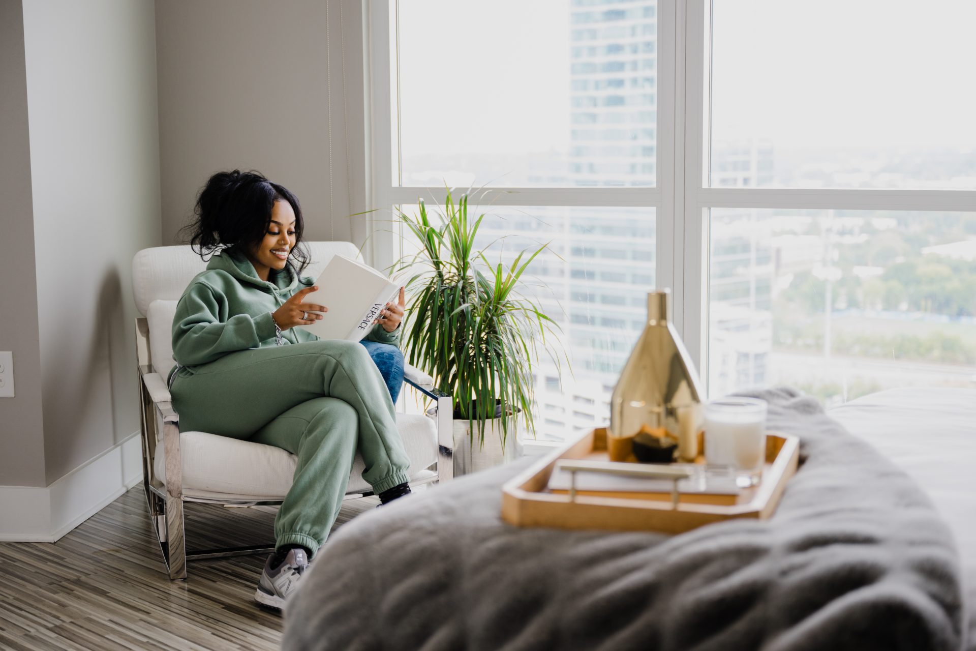 Woman in a green outfit relaxing in a white accent chair reading a book beside large windows with natural light for a luxury apartment lifestyle brand shoot