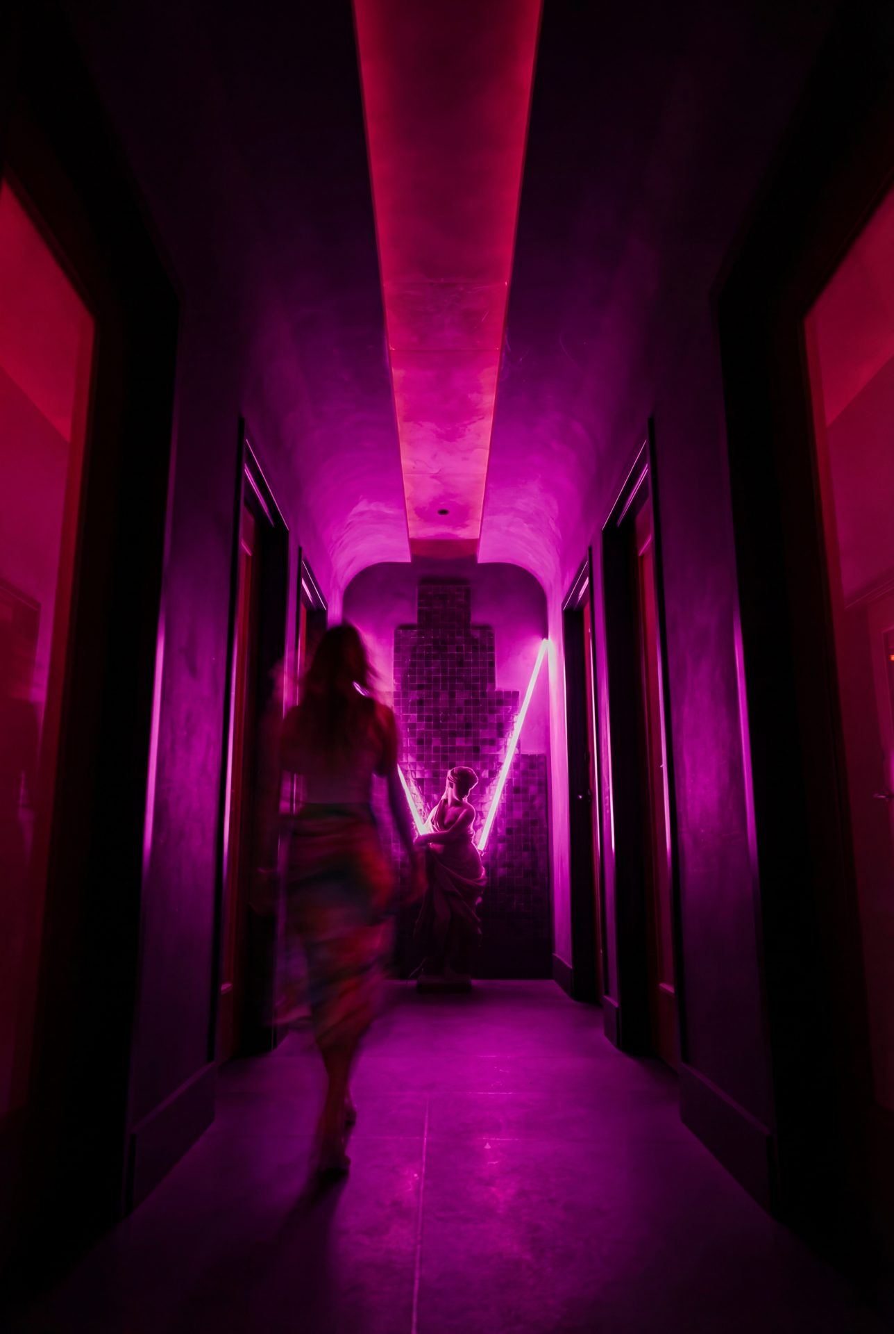 Person walking through a narrow corridor bathed in vibrant pink and purple neon lighting creating a dramatic moody atmosphere
