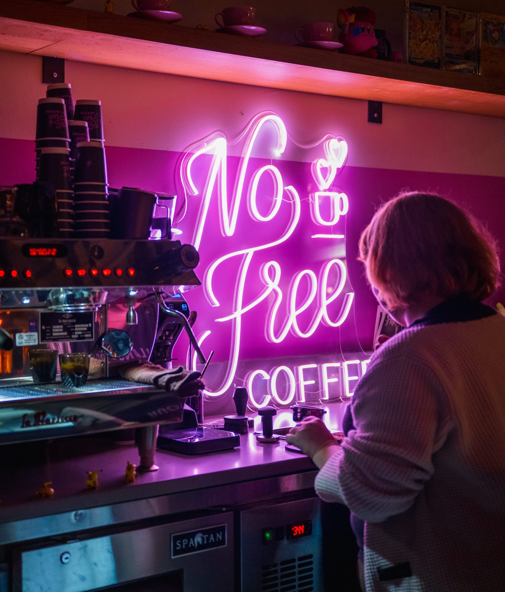 Bright pink neon sign reading No Free Coffee mounted on a wall above a coffee bar counter with a barista working nearby