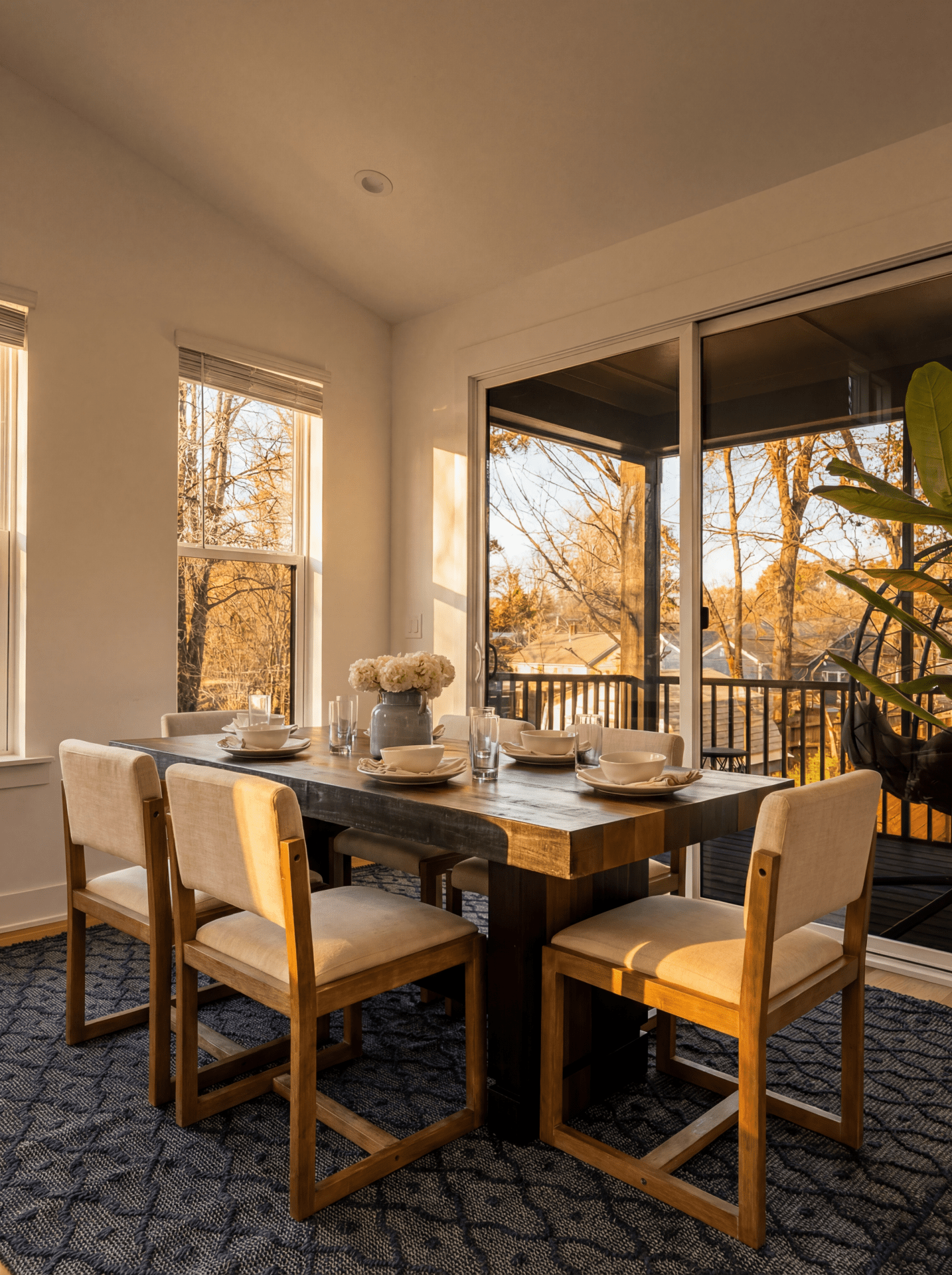 Sunlit dining room with a wooden table, upholstered chairs, and large windows overlooking autumn trees